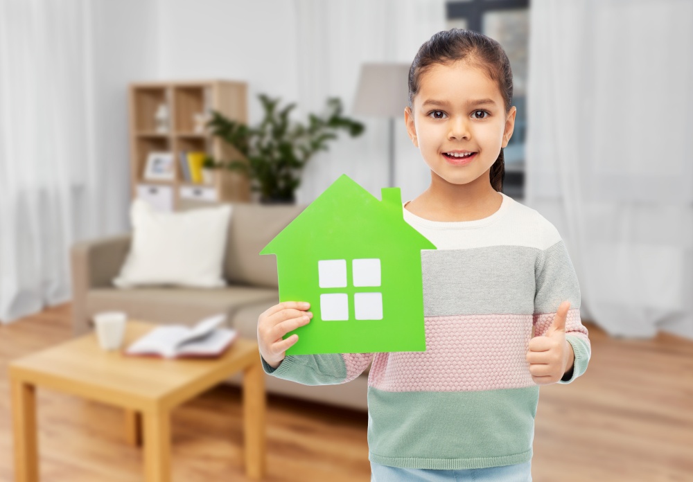 eco living, environment and sustainability concept - smiling little girl holding green house icon showing thumbs up over home background. smiling little girl with green house icon at home