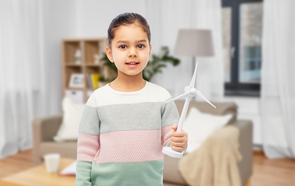 sustainable energy, power and people concept - happy smiling girl with toy wind turbine over home background. smiling girl with toy wind turbine at home