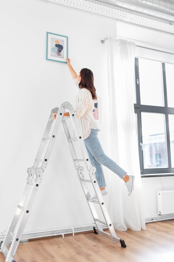 home improvement, decoration and people concept - happy smiling woman on ladder decorating home with art. woman on ladder decorating home with art