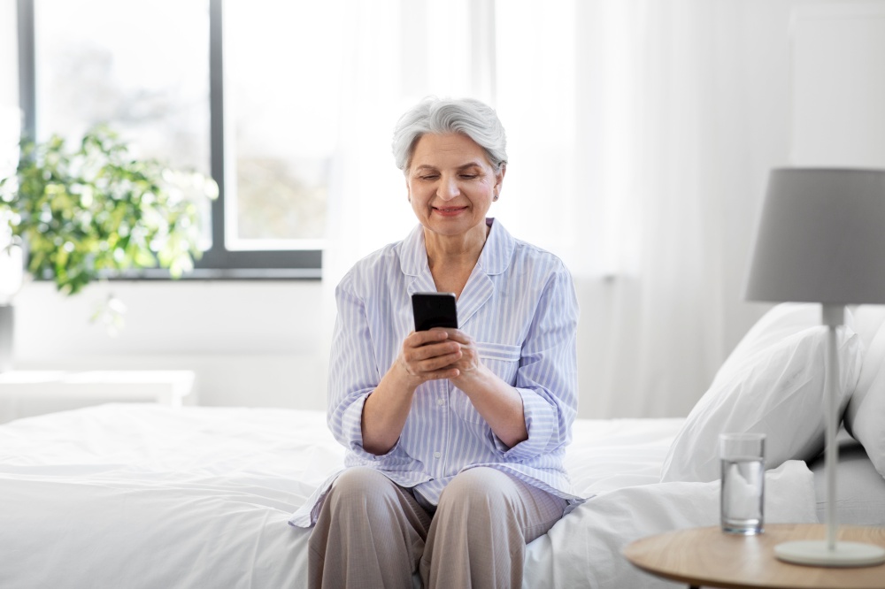 old age, technology and people concept - happy smiling senior woman in pajamas with smartphone sitting on bed at home bedroom. happy senior woman with smartphone on bed at home