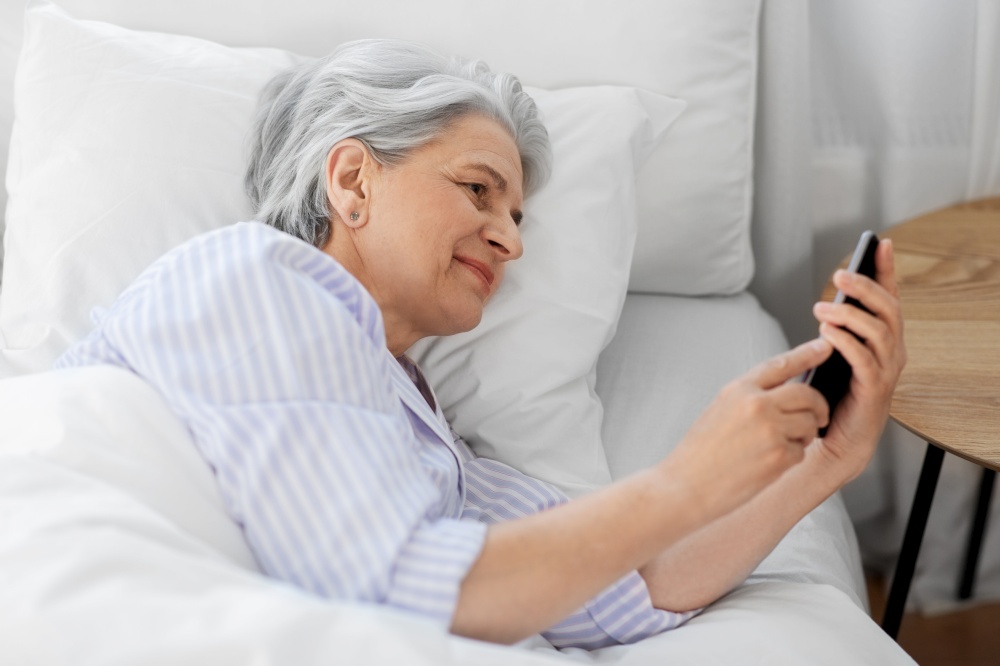 old age and people concept - happy smiling senior woman with smartphone lying in bed at home bedroom. happy senior woman with smartphone in bed at home