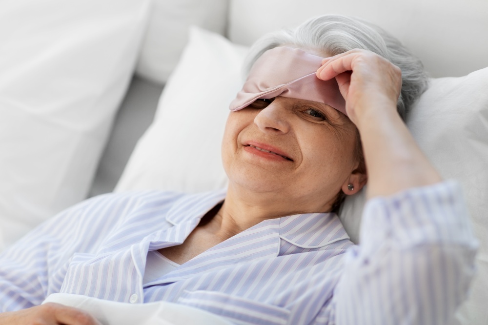 old age and people concept - happy smiling senior woman with eye sleeping mask in bed at home bedroom. senior woman with eye sleeping mask in bed at home