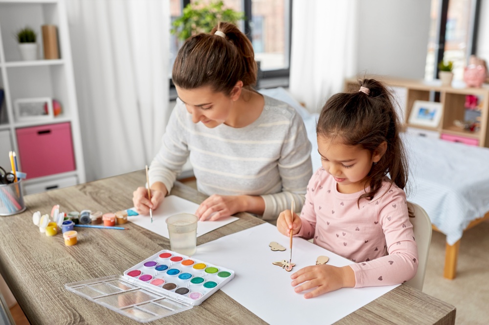 family, motherhood and leisure concept - mother spending time with her little daughter with colors painting chipboard items at home. mother with little daughter drawing at home