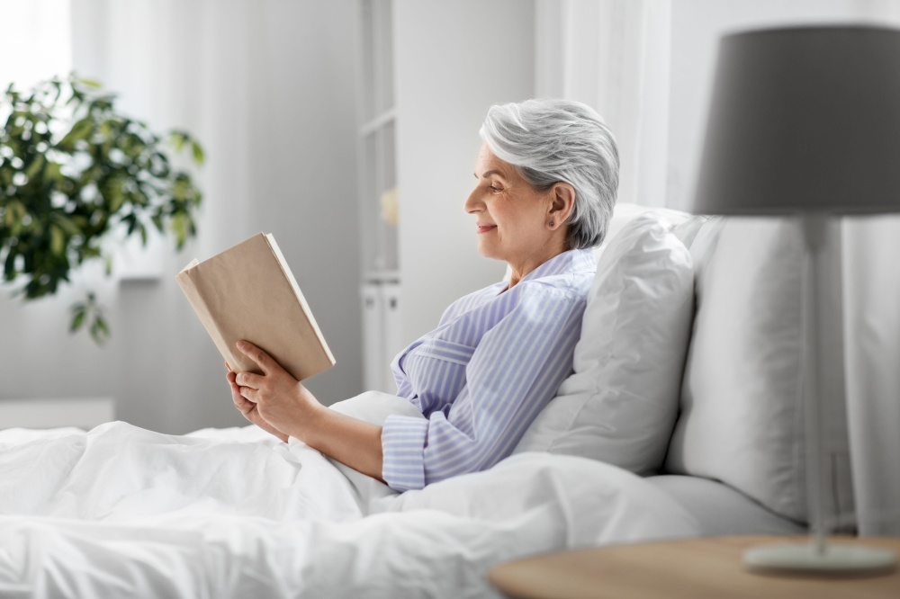 technology, old age and people concept - senior woman reading book in bed at home bedroom. senior woman reading book in bed at home bedroom