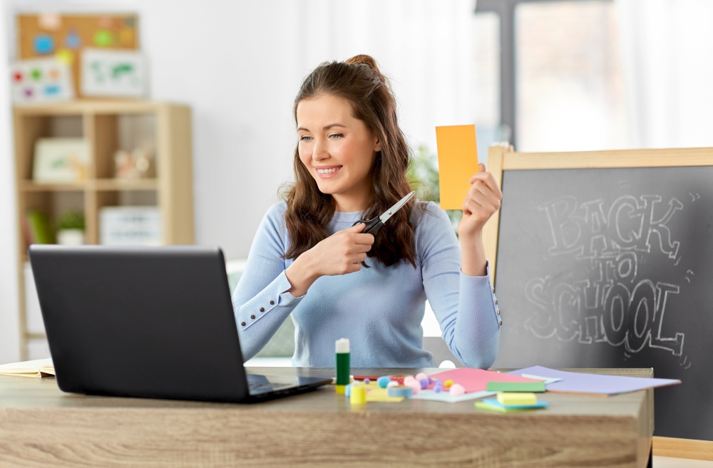 distant education, school and people concept - happy smiling female teacher with laptop computer, color paper and scissors having online class of arts and crafts at home. teacher having online class of arts and crafts