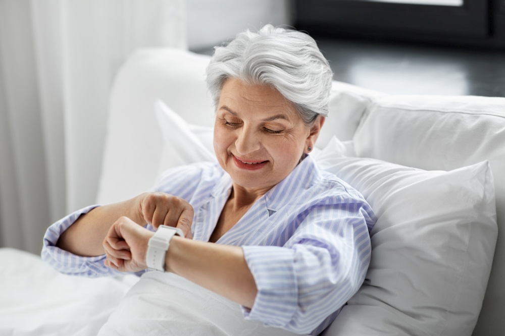 old age and people concept - happy smiling senior woman in pajamas with smart watch sitting in bed at home bedroom. happy senior woman sitting in bed at home bedroom
