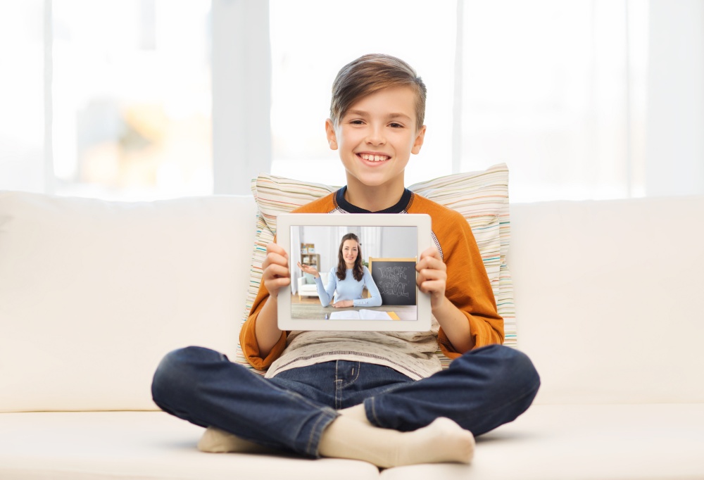 online education, technology and children concept - smiling boy sitting on couch at home and holding tablet pc computer with teacher on screen. happy boy with teacher on tablet computer at home