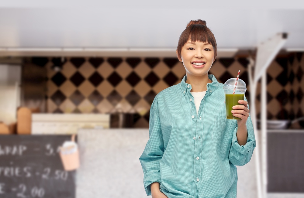 diet, healthy eating and detox concept - happy smiling young asian woman drinking green vegetable juice or smoothie from plastic cup with paper straw over food truck background. happy asian woman with juice over food truck