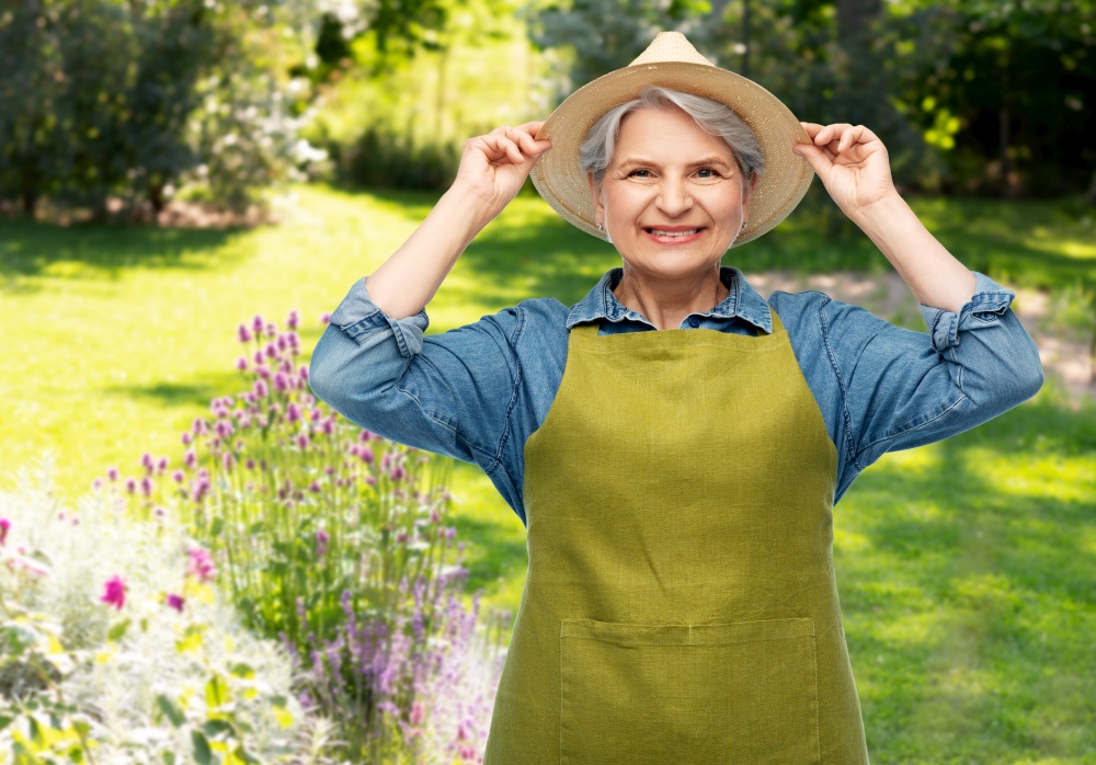 gardening, farming and old people concept - portrait of smiling senior woman in green apron and straw hat over summer garden background. portrait of smiling senior woman in garden apron