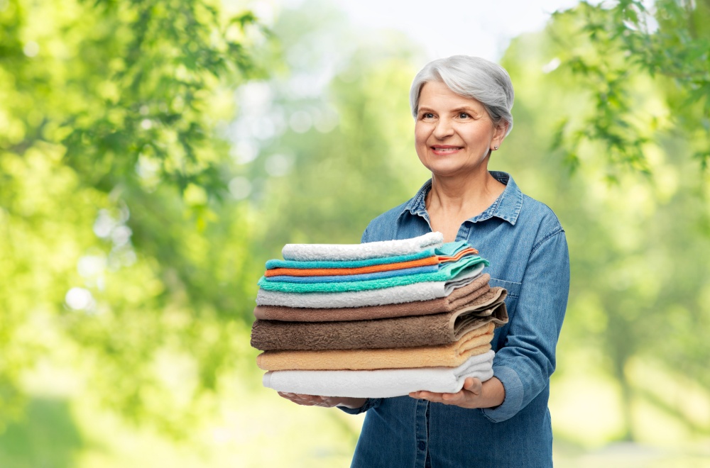 cleaning, laundry and old people concept - portrait of smiling senior woman in denim shirt with pile of clean and folded bath towels over green natural background. smiling senior woman with clean bath towels