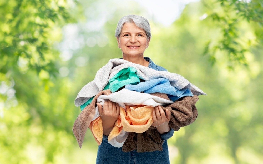 cleaning, laundry and old people concept - portrait of smiling senior woman in denim shirt with heap of bath towels over green natural background. smiling senior woman with heap of bath towels