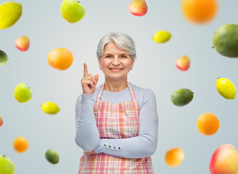 cooking, culinary and old people concept - portrait of smiling senior woman in kitchen apron pointing finger up over fruits on grey background. smiling senior woman in apron pointing finger up