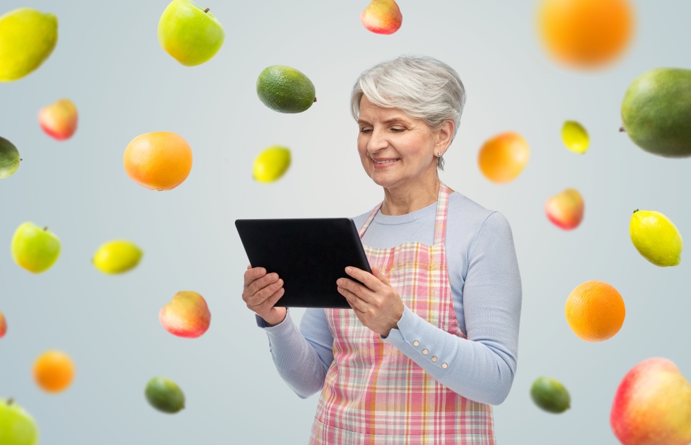 cooking, culinary and old people concept - happy smiling senior woman in kitchen apron with tablet pc computer over fruits on grey background. smiling senior woman in apron with tablet computer