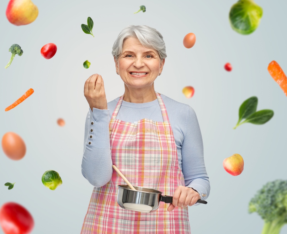 food cooking, culinary and old people concept - portrait of smiling senior woman in kitchen apron with pot and spoon over fruits on grey background. senior woman in apron with pot cooking food