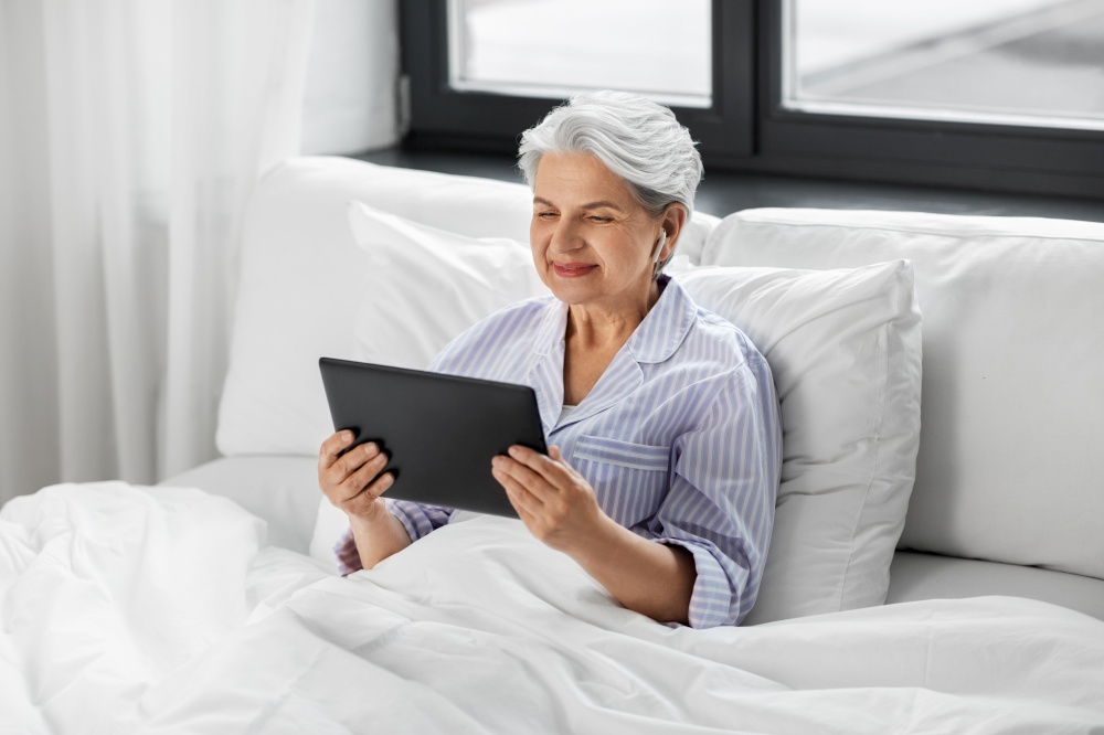 technology, old age and people concept - senior woman with tablet pc computer and wireless earphones in bed at home bedroom. senior woman with tablet pc and earphones in bed