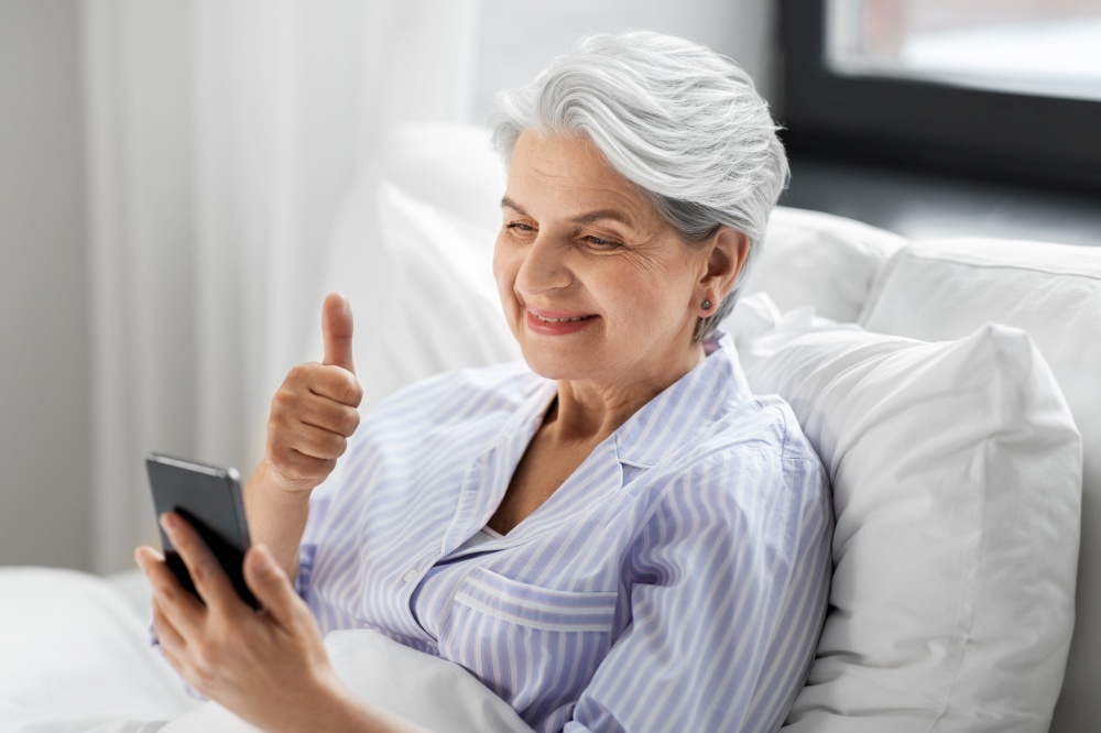 old age, technology and people concept - happy smiling senior woman in pajamas using smartphone sitting in bed and having video call and showing thumbs up at home bedroom. senior woman with phone having video call in bed