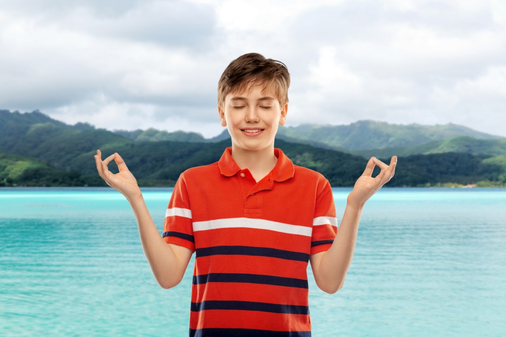 childhood, fashion and people concept - happy smiling boy in red polo t-shirt meditating over hills and ocean background in french polynesia. happy smiling meditating boy over hills and ocean