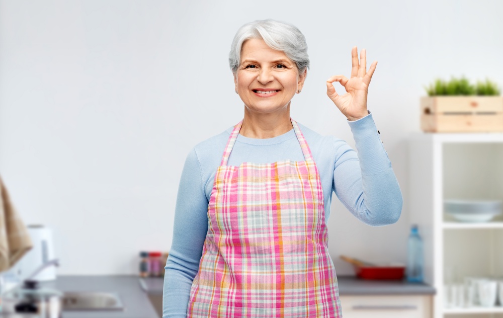 cooking, culinary and old people concept - portrait of smiling senior woman in apron showing ok hand sign over home kitchen background. smiling senior woman showing ok gesture at kitchen