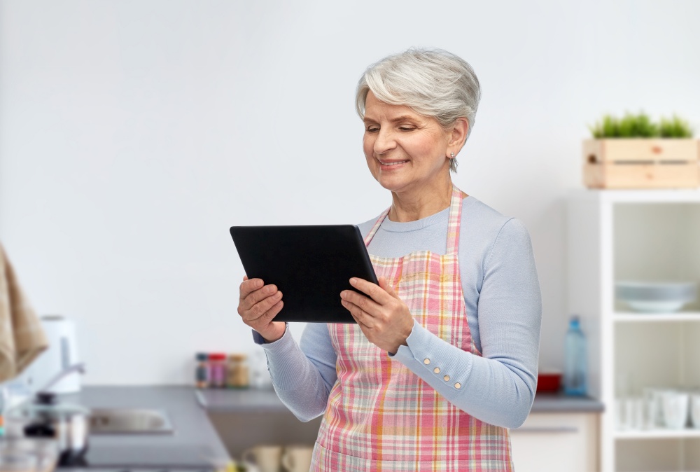 cooking, culinary and old people concept - happy smiling senior woman in apron with tablet pc computer over home kitchen background. smiling senior woman with tablet pc at kitchen