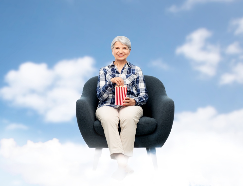 leisure, entertainment and old people concept - smiling senior woman sitting in modern armchair and eating popcorn from striped bucket over blue sky and clouds background. senior woman eating popcorn sitting in armchair