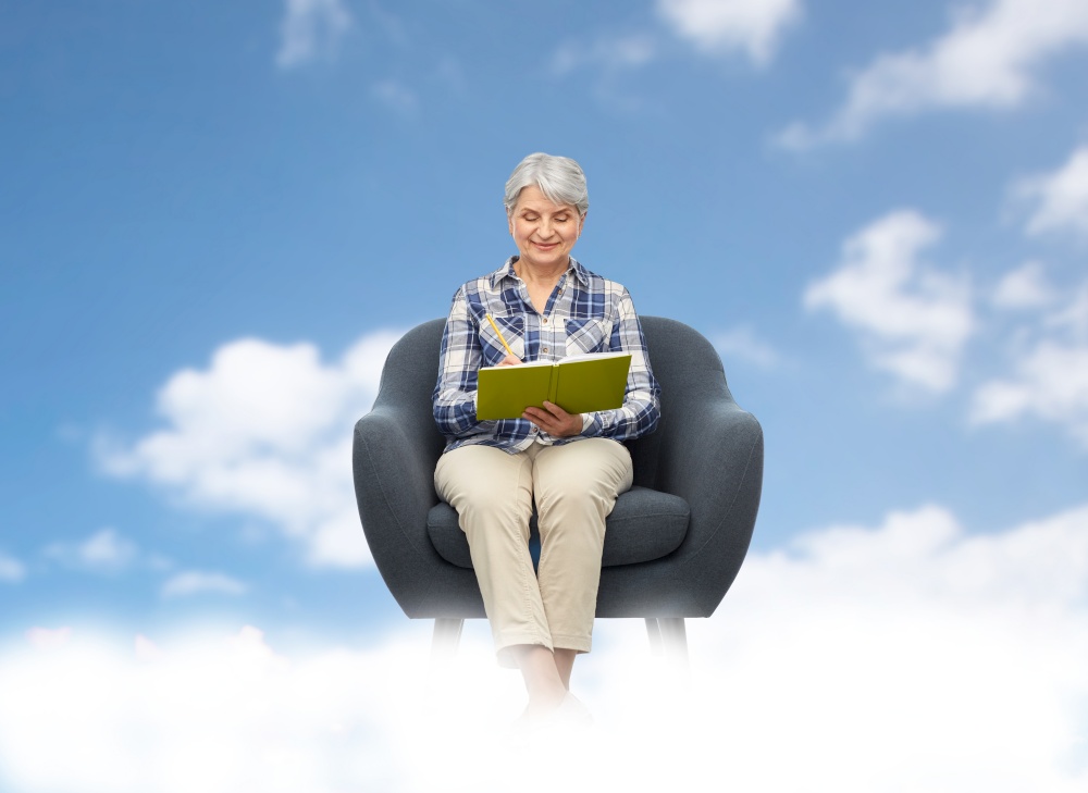 inspiration, planning and old people concept - smiling senior woman sitting in modern armchair and writing to diary over blue sky and clouds background. smiling senior woman in armchair writing to diary