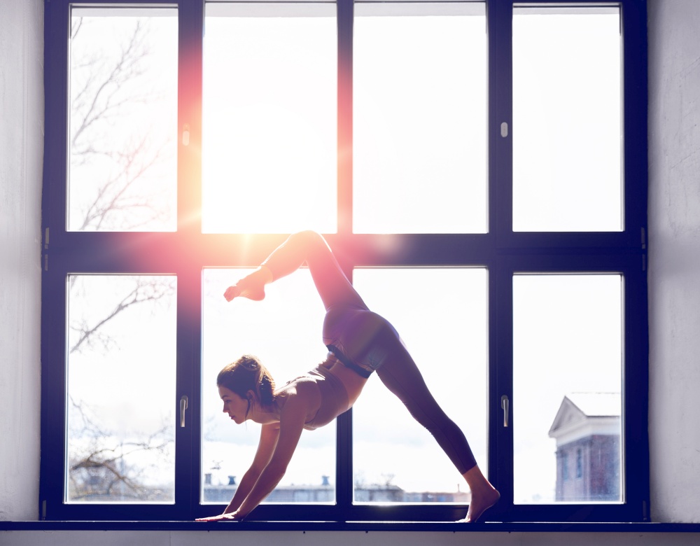fitness, sport and healthy lifestyle concept - woman doing yoga exercise on window sill at studio over sunlight. woman doing yoga exercise on window sill at studio