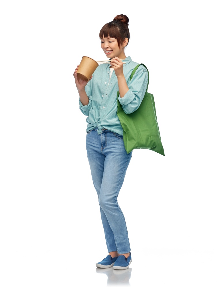 sustainability, food shopping and eco friendly concept - asian woman in turquoise shirt and jeans holding reusable green tote bag eating takeaway wok with chopsticks over white background. asian woman with reusable bag for food and wok