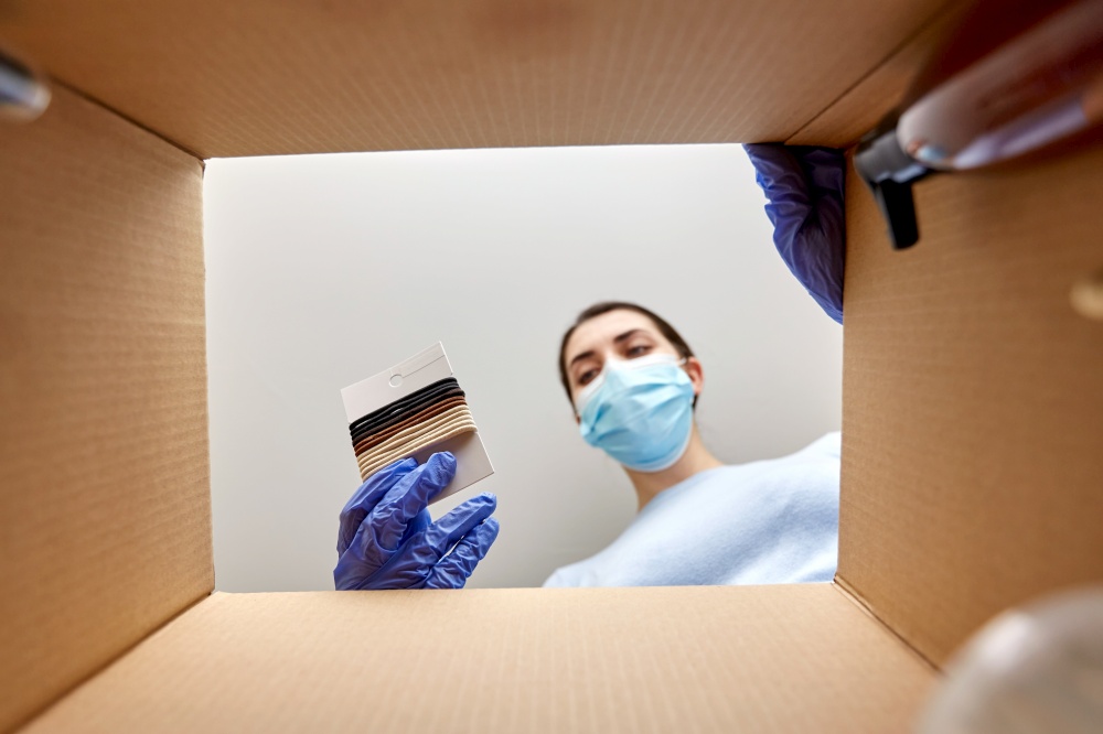 home delivery, shipping and pandemic concept - woman in protective medical mask and gloves unpacking parcel box with elastic hair bands and beauty products. woman in mask unpacking parcel box with hair bands