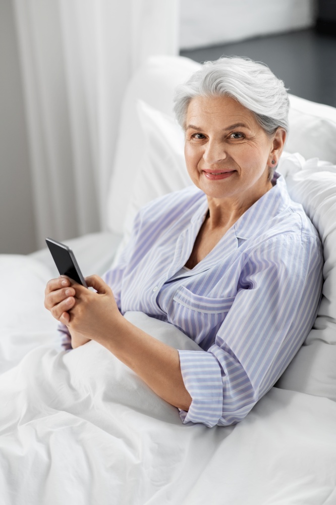 old age, technology and people concept - happy smiling senior woman in pajamas using smartphone sitting in bed at home bedroom. happy senior woman using smartphone in bed at home