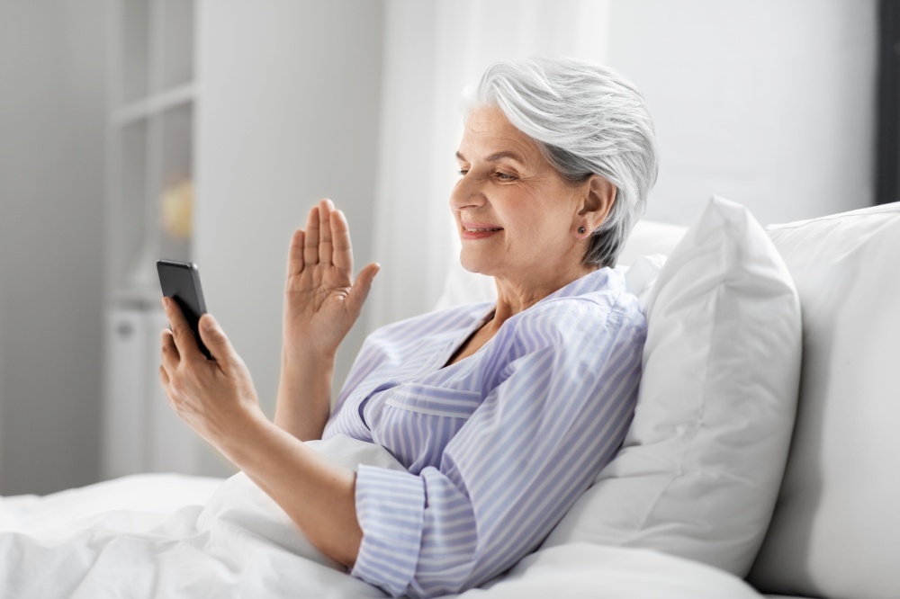 old age, technology and people concept - happy smiling senior woman in pajamas using smartphone sitting in bed and having video call at home bedroom. senior woman with phone having video call in bed