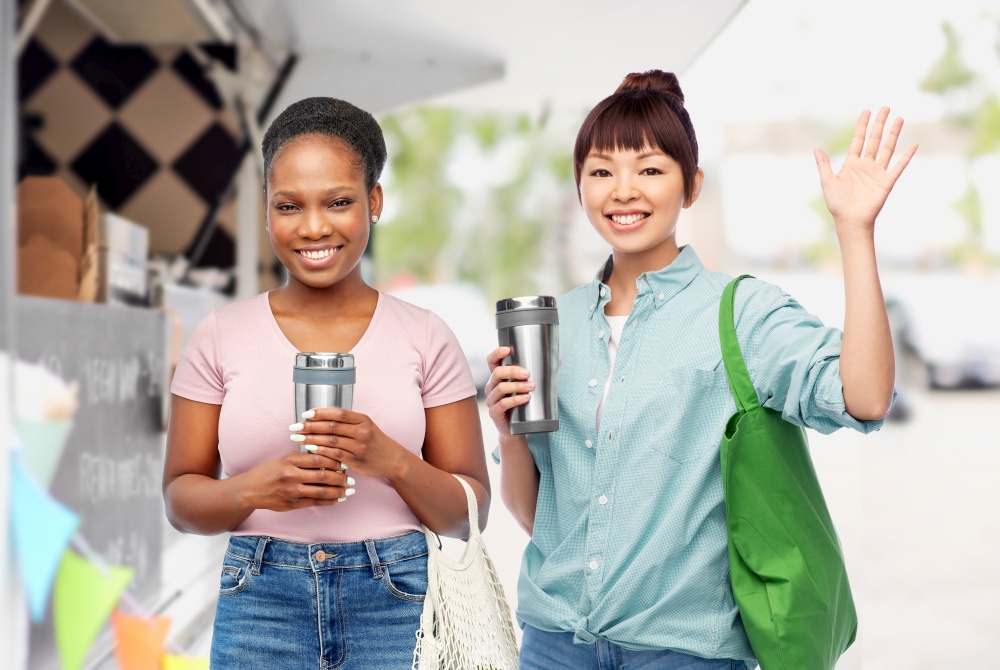sustainability, eco living and people concept - asian and african american women with thermo cups for hot drinks and reusable bags for food shopping waving hand over food truck background. women with tumblers and bags over food truck