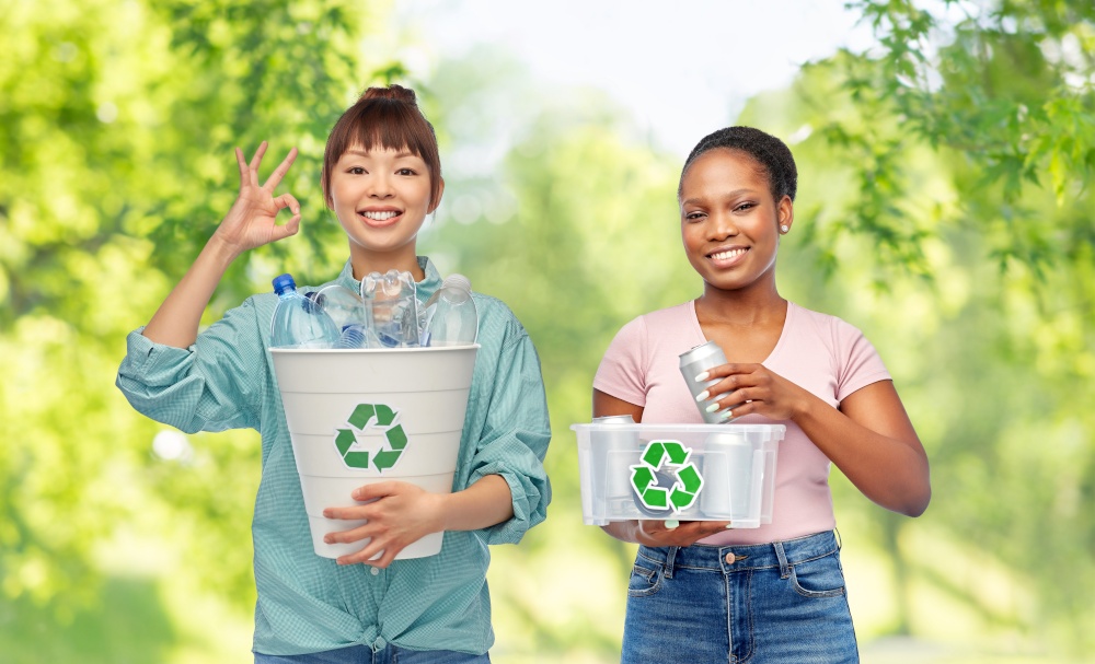 metal recycling, waste sorting and sustainability concept - happy asian and african american women holding boxes with plastic bottles and tin cans over green natural background. happy women sorting plastic and metallic waste