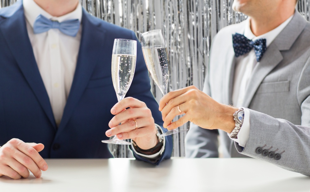 homosexuality, same-sex marriage and lgbt concept - close up of married male gay couple in suits and bow-ties drinking sparkling wine and clinking glasses on wedding over foil curtain on background. close up of male gay couple with champagne glasses