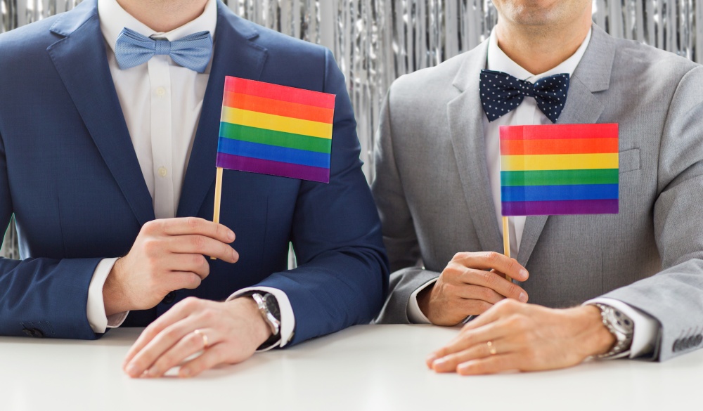 homosexuality, same-sex marriage and lgbt concept - close up of happy male gay couple in suits and bow-ties with wedding rings holding rainbow flags over foil party curtain on background. close up of male gay couple holding rainbow flags