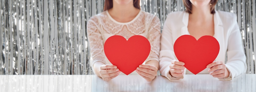 homosexuality, valentine&rsquo;s day and love concept - close up of happy lesbian couple holding red paper hearts over foil party curtain on background. close up of happy lesbian couple with red hearts
