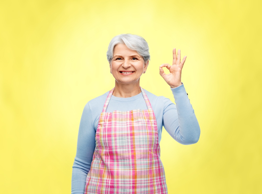cooking, culinary and old people concept - portrait of smiling senior woman in kitchen apron showing ok hand sign over illuminating yellow background. smiling senior woman in apron showing ok gesture