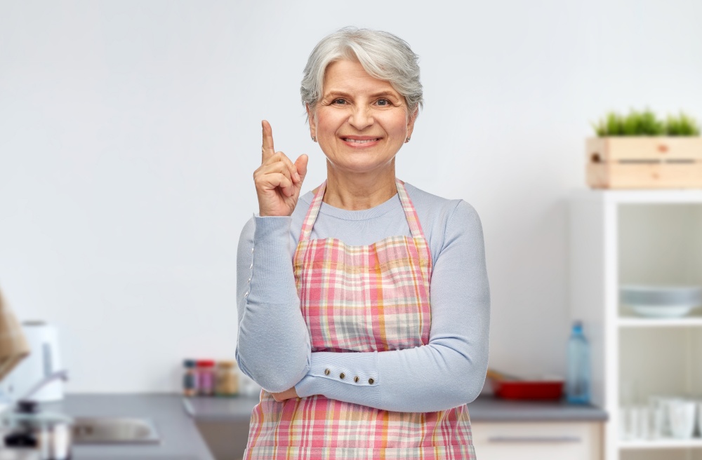 cooking, culinary and old people concept - portrait of smiling senior woman in apron pointing finger up over home kitchen background. smiling senior woman pointing finger up at kitchen