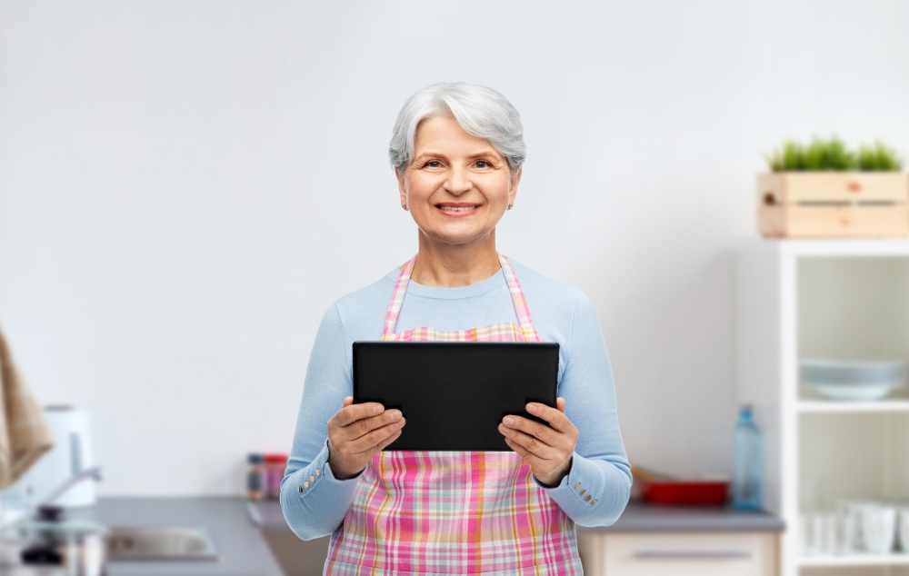 cooking, culinary and old people concept - portrait of smiling senior woman r in apron with tablet pc compute over home kitchen background. smiling senior woman with tablet pc at kitchen
