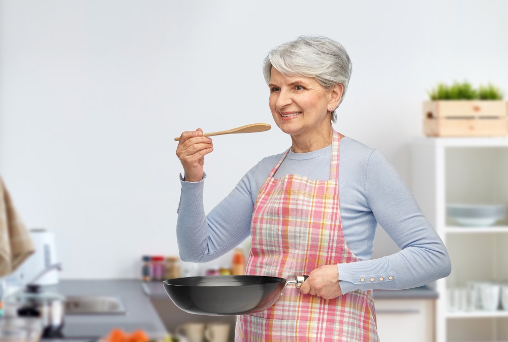 cooking, culinary and old people concept - portrait of smiling senior woman in apron with frying pan and spoon tasting food over home kitchen background. smiling senior woman with frying pan at kitchen