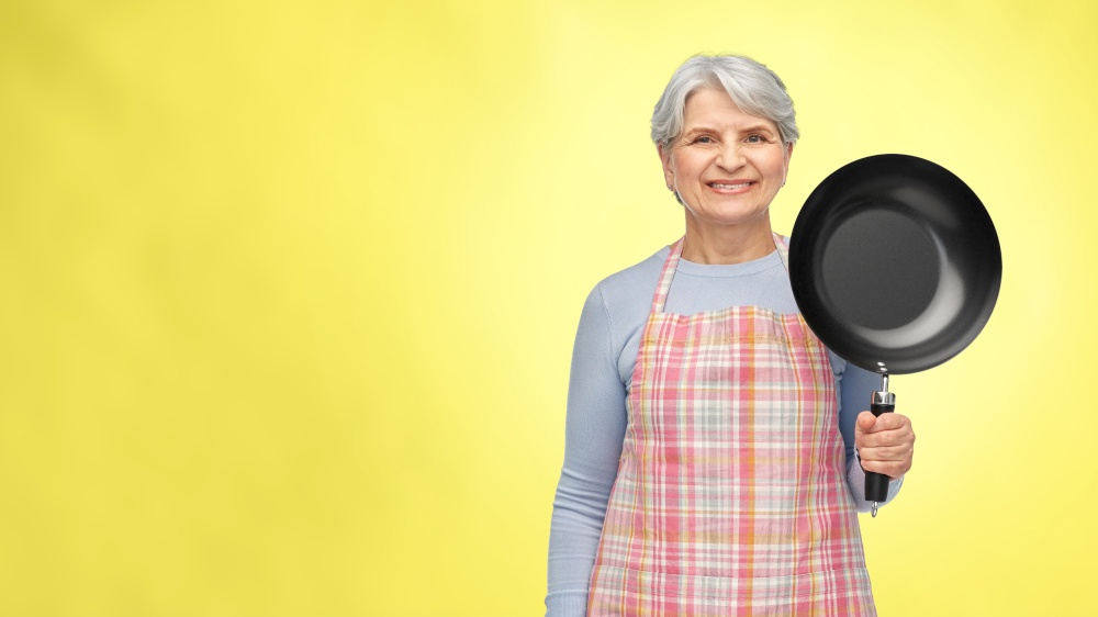 food cooking, culinary and old people concept - portrait of smiling senior woman in kitchen apron with frying pan over illuminating yellow background. smiling senior woman in apron with frying pan