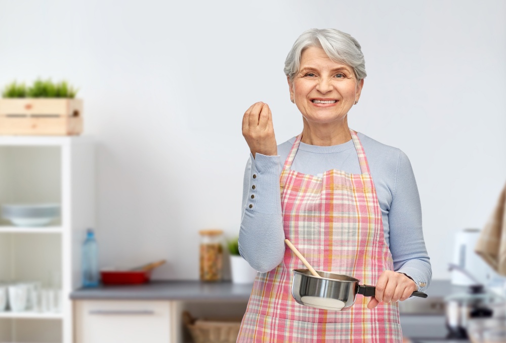 food cooking, culinary and old people concept - portrait of smiling senior woman in apron with pot and spoon over home kitchen background. senior woman with pot cooking food at kitchen