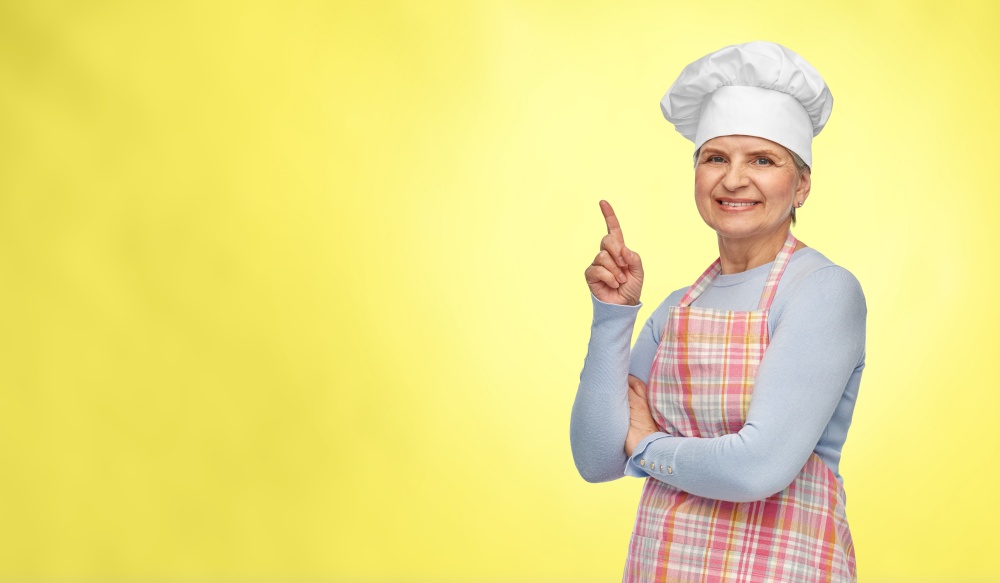 cooking, culinary and old people concept - portrait of smiling senior woman or chef in toque in kitchen apron pointing finger up over illuminating yellow background. smiling senior woman or chef pointing finger up