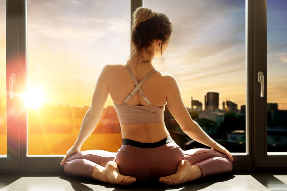 fitness, sport and healthy lifestyle concept - woman doing yoga exercise on window sill at studio over city sunset or sunrise on background. woman doing yoga exercise on window sill at studio