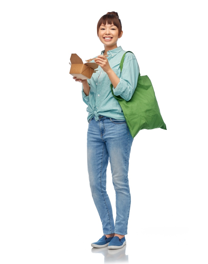 sustainability, food shopping and eco friendly concept - happy smiling asian woman in turquoise shirt and jeans holding reusable green tote bag eating takeaway wok over white background. asian woman with reusable bag for food and wok