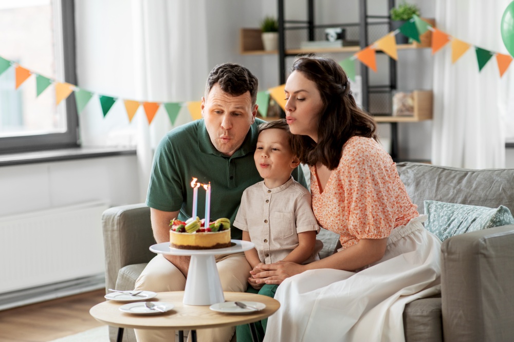 family, holidays and people concept - portrait of happy mother, father and little son blowing together to four candles burning on birthday cake sitting on sofa at home party. happy family with birthday cake at home