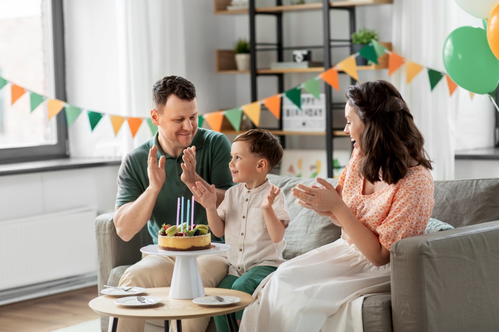 family, holidays and people concept - portrait of happy mother, father and little son with four candles on birthday cake clapping hands at home party party. happy family with birthday cake at home