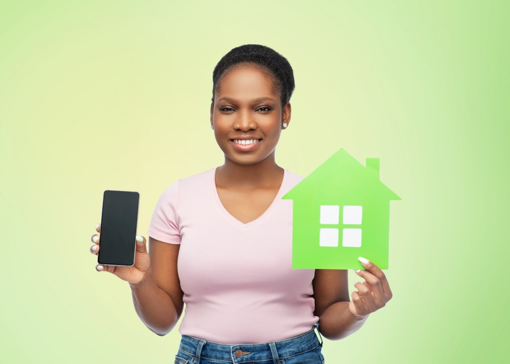 eco living, environment and sustainability concept - happy smiling young african american woman holding smartphone and green house over natural background. african woman with smartphone and green house