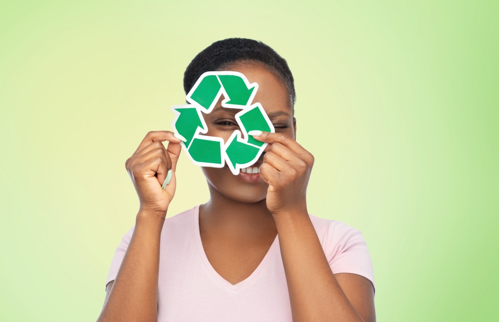 eco living, environment and sustainability concept - portrait of happy smiling young african american woman looking through recycling sign over green background. smiling asian woman holding green recycling sign