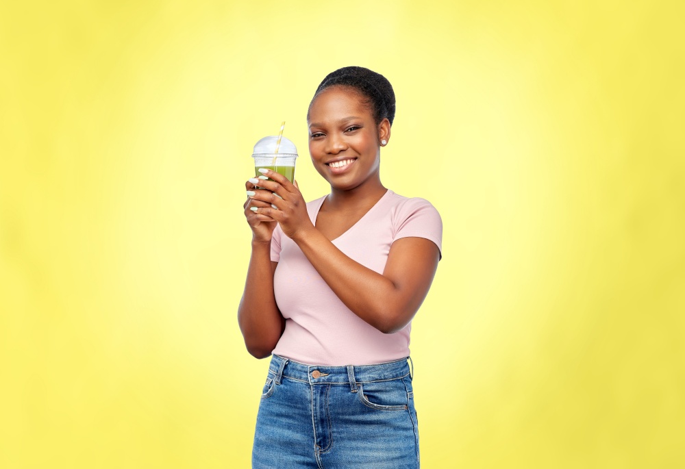 diet, healthy eating and detox concept - happy smiling young african american woman drinking green vegetable juice or smoothie from plastic cup with paper straw over illuminating yellow background. happy african american woman drinking green juice