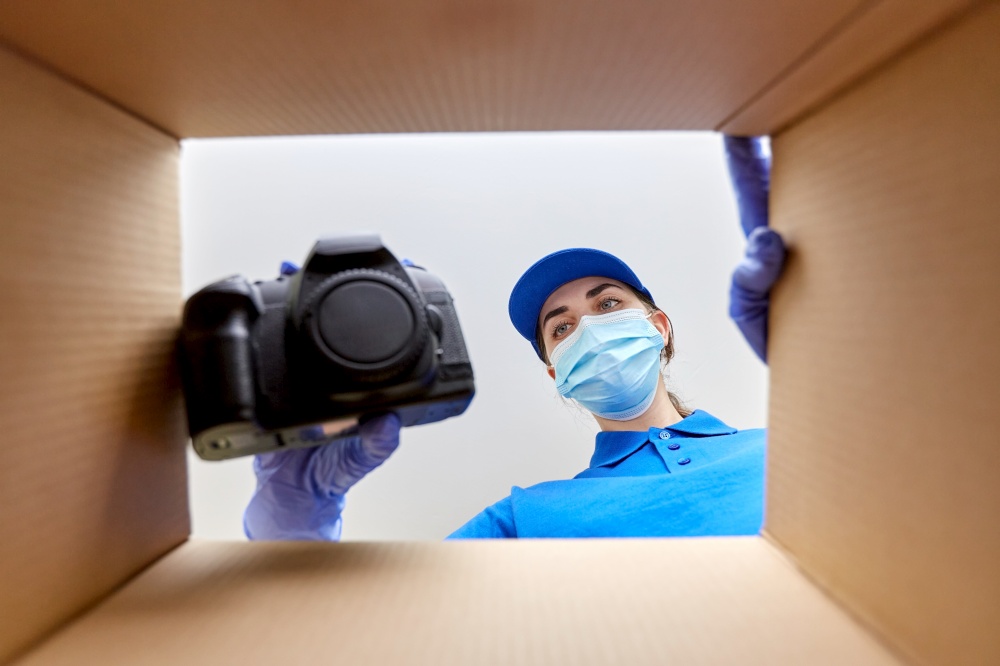 delivery, shipping and pandemic concept - woman in protective medical mask and gloves packing digital camera into parcel box with foam peanuts. woman in mask packing camera into parcel box
